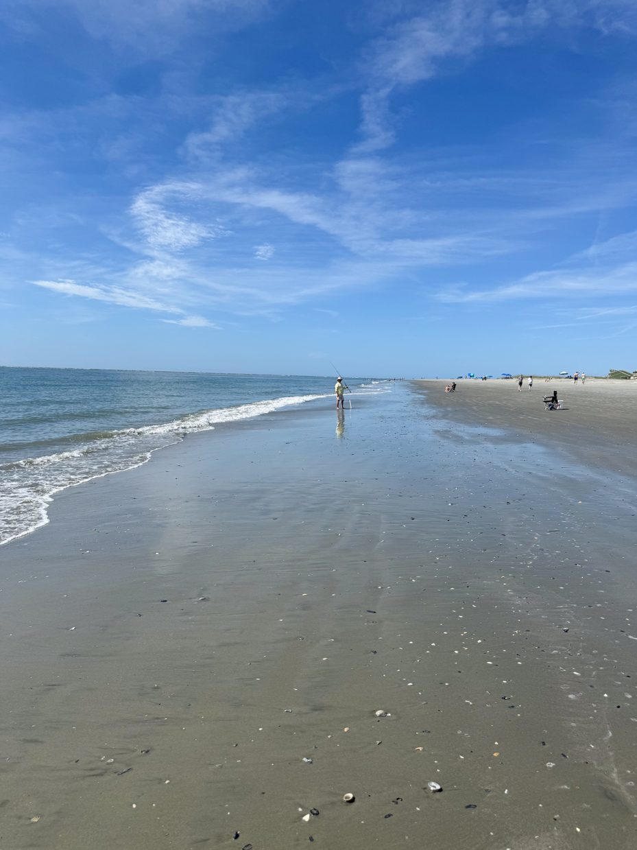 man-fishing-along-a-peaceful-sandy-beach-under-a-bright-blue-sky-with-a-few-scattered-clouds