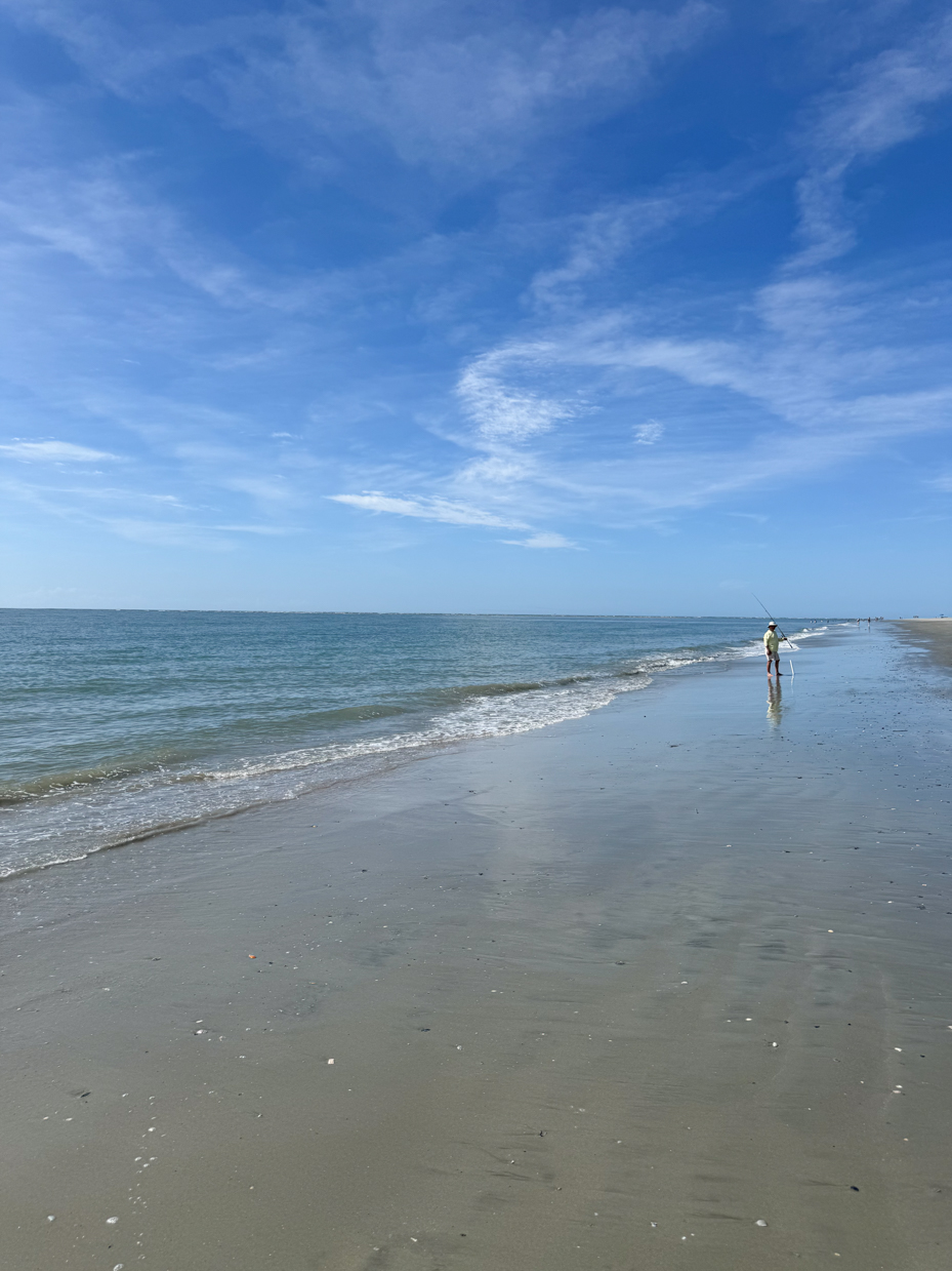 man-fishing-on-a-quiet-beach-with-gentle-waves-under-a-clear-blue-sky-with-wispy-clouds