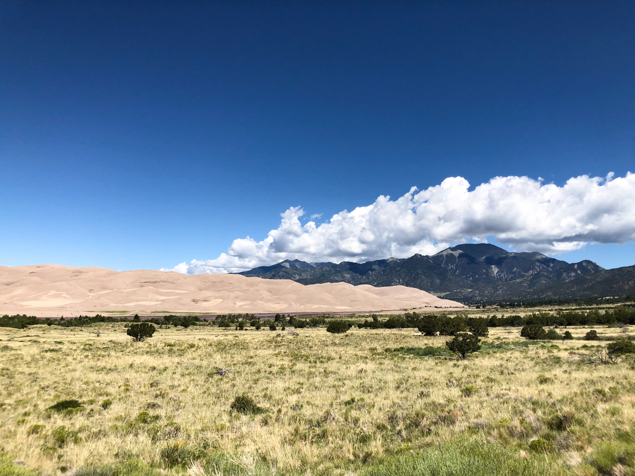 expansive-sand-dunes-with-mountain-range-and-open-grassland-under-bright-blue-skies