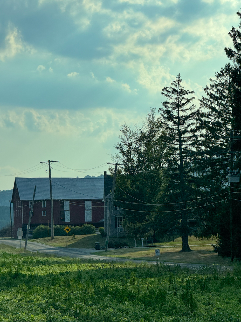 a-quiet-rural-road-weaves-past-a-red-barn-and-a-cluster-of-tall-pine-trees-with-power-lines-stretching-overhead-framed-by-a-dramatic-cloudy-sky-and-green-fields-in-the-foreground-capturing-the-calm