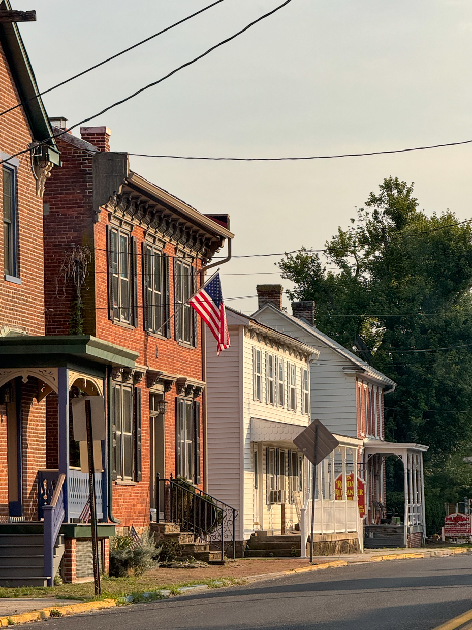 an-american-flag-proudly-waves-from-the-porch-of-a-charming-red-brick-house-in-a-quaint-tree-lined-town-with-a-row-of-historic-homes-displaying-their-cozy-porches-along-a-peaceful-street