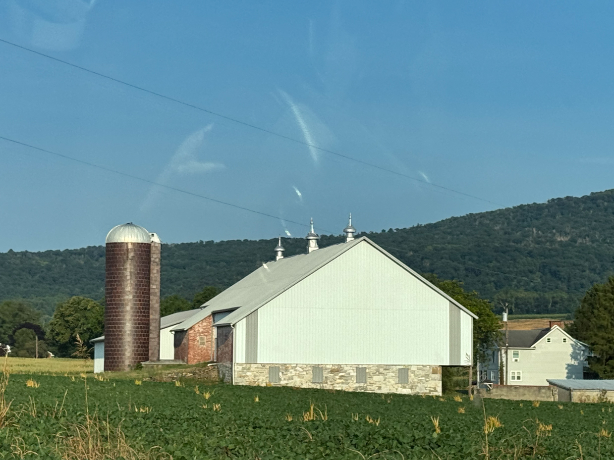 farm-scene-with-a-barn-featuring-a-metal-roof-and-brick-silo-surrounded-by-green-crops-and-backed-by-a-mountain-range-under-a-clear-blue-sky-in-late-afternoon-light