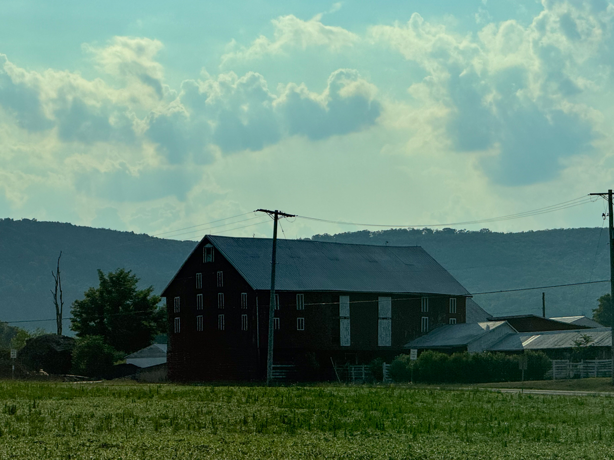 rustic-barn-standing-against-a-backdrop-of-rolling-hills-shrouded-in-moody-clouds-with-telephone-wires-cutting-across-the-sky-and-a-field-in-the-foreground-creating-a-serene-and-quiet-farm-scene