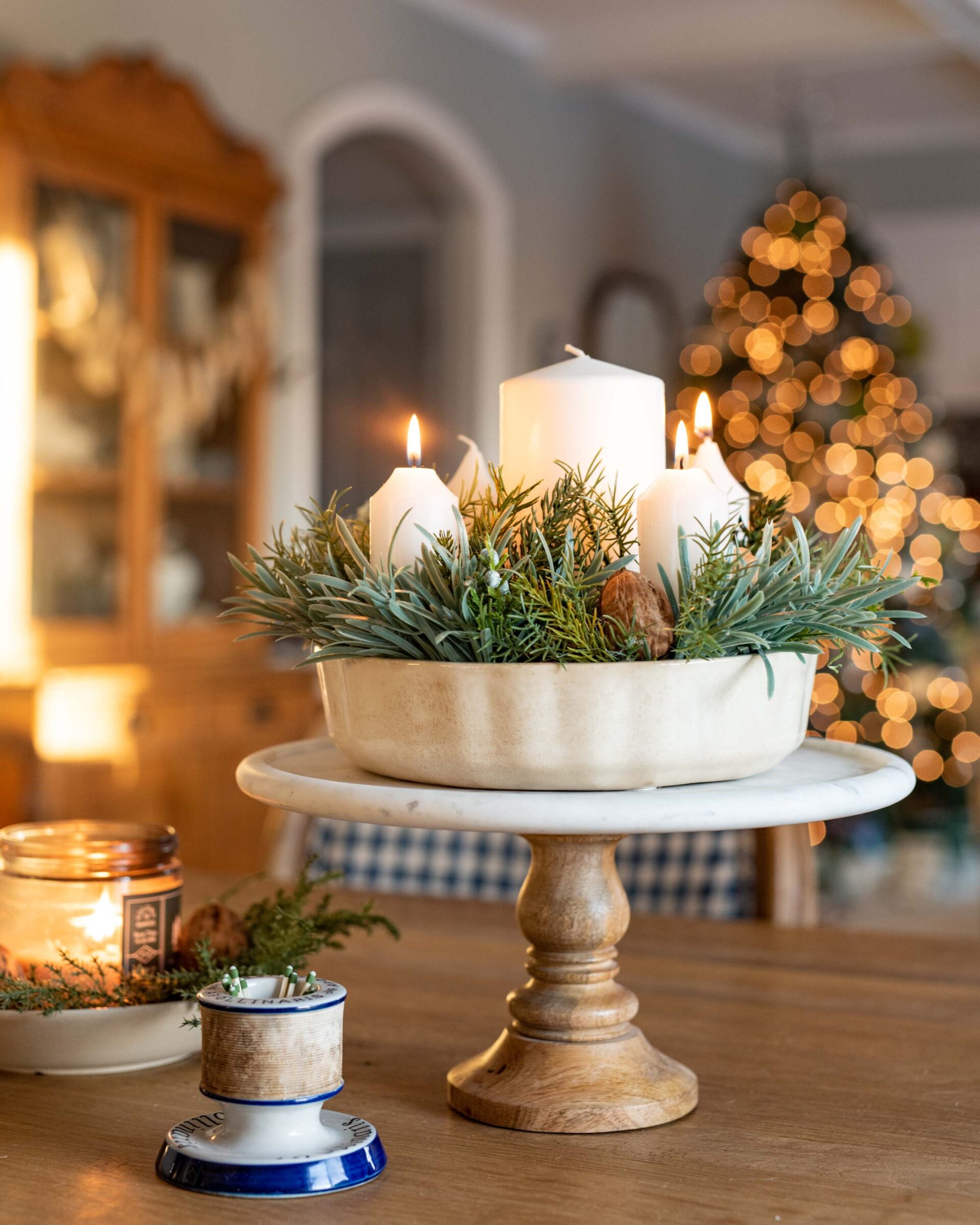 cozy-christmas-centerpiece-with-white-pillar-candles-fresh-greenery-and-wooden-pedestal-riser-with-bokeh-tree-lights-in-rustic-home-interior