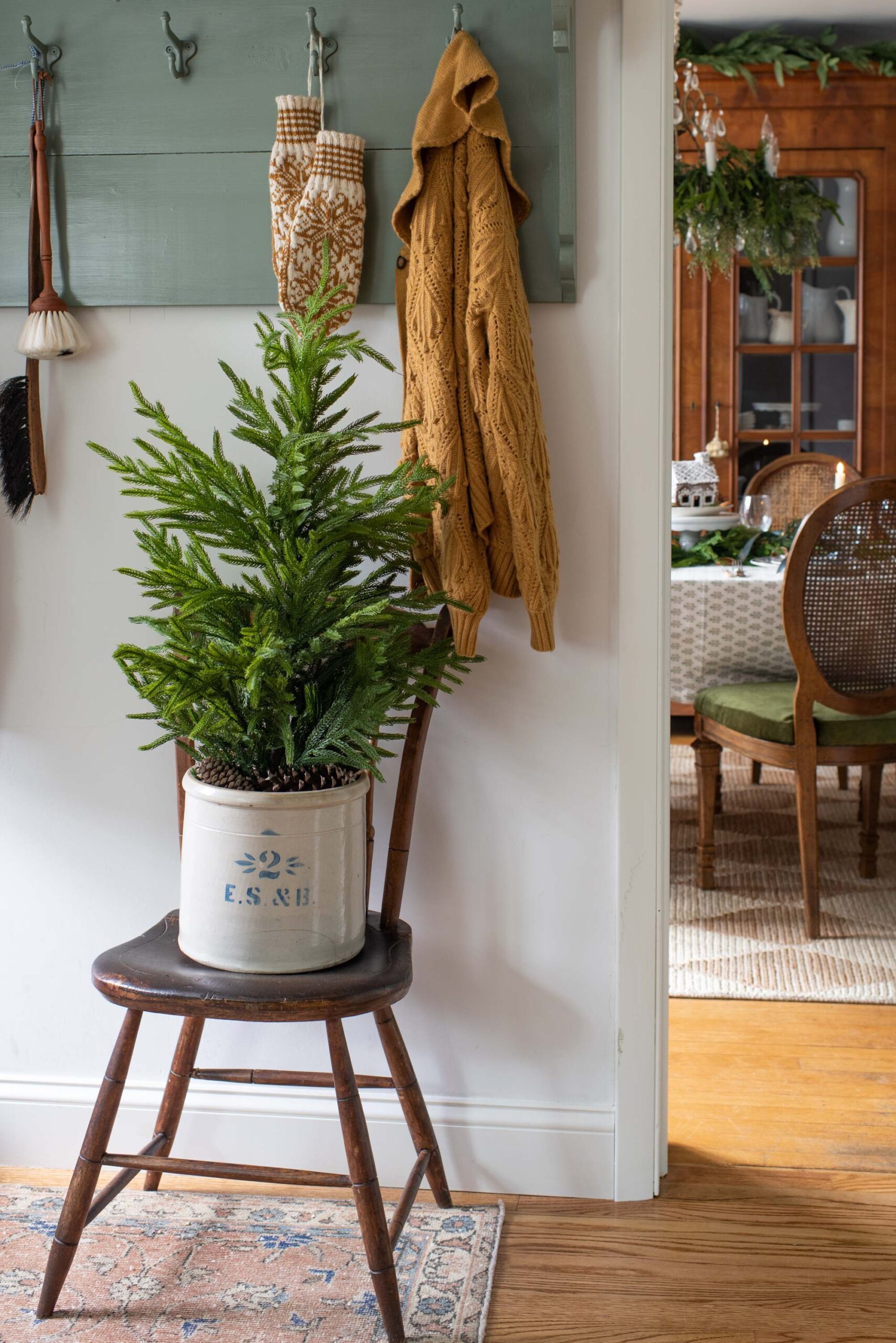 Cozy holiday entryway with evergreen plant in vintage crock on wooden stool, knit cardigan hanging from hook, and dining area in background artist reference photo