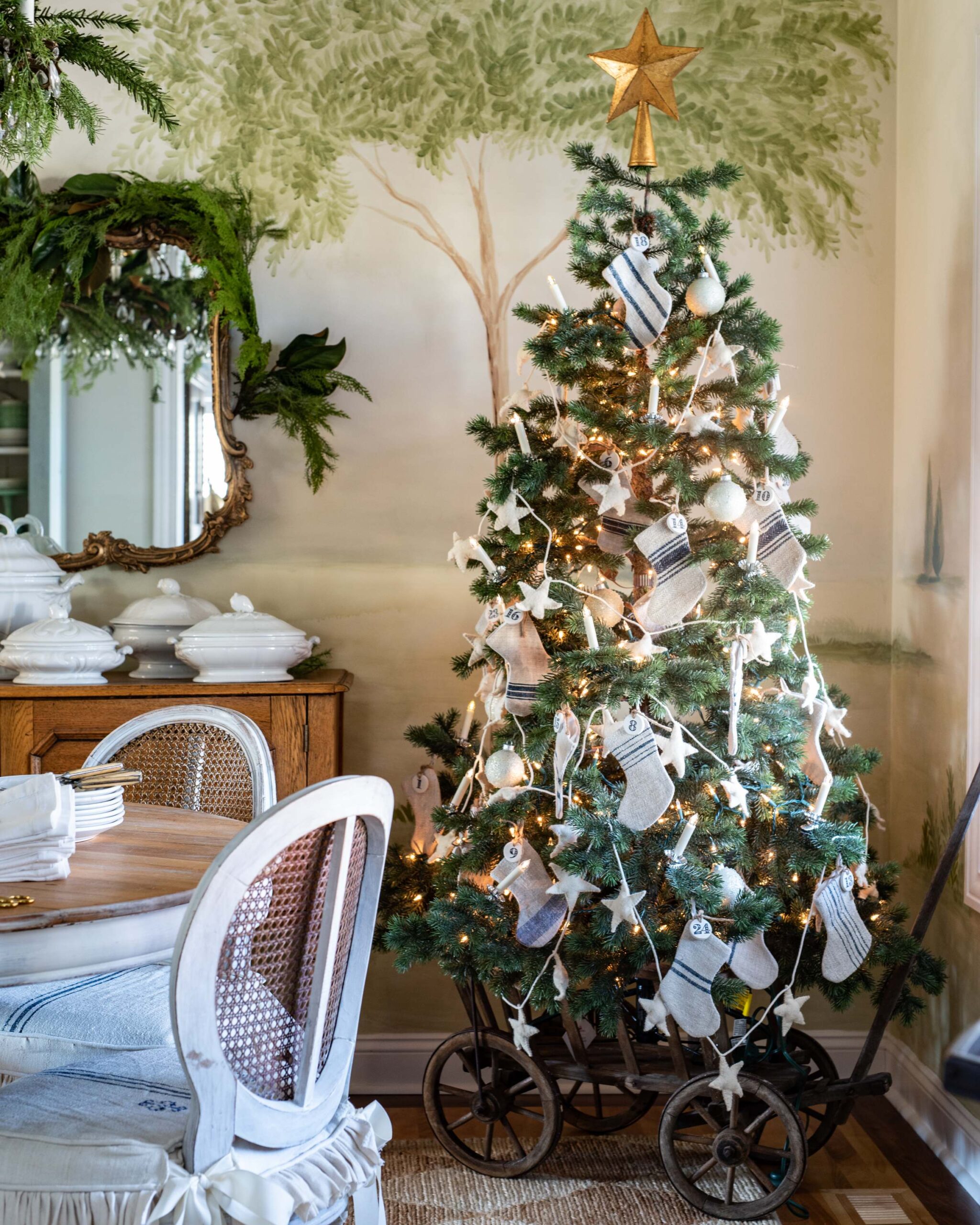 Festive Christmas tree with white and grey mitten ornaments and gold star topper, next to vintage wood cart with white linen chair covers in cozy dining room artist reference photo