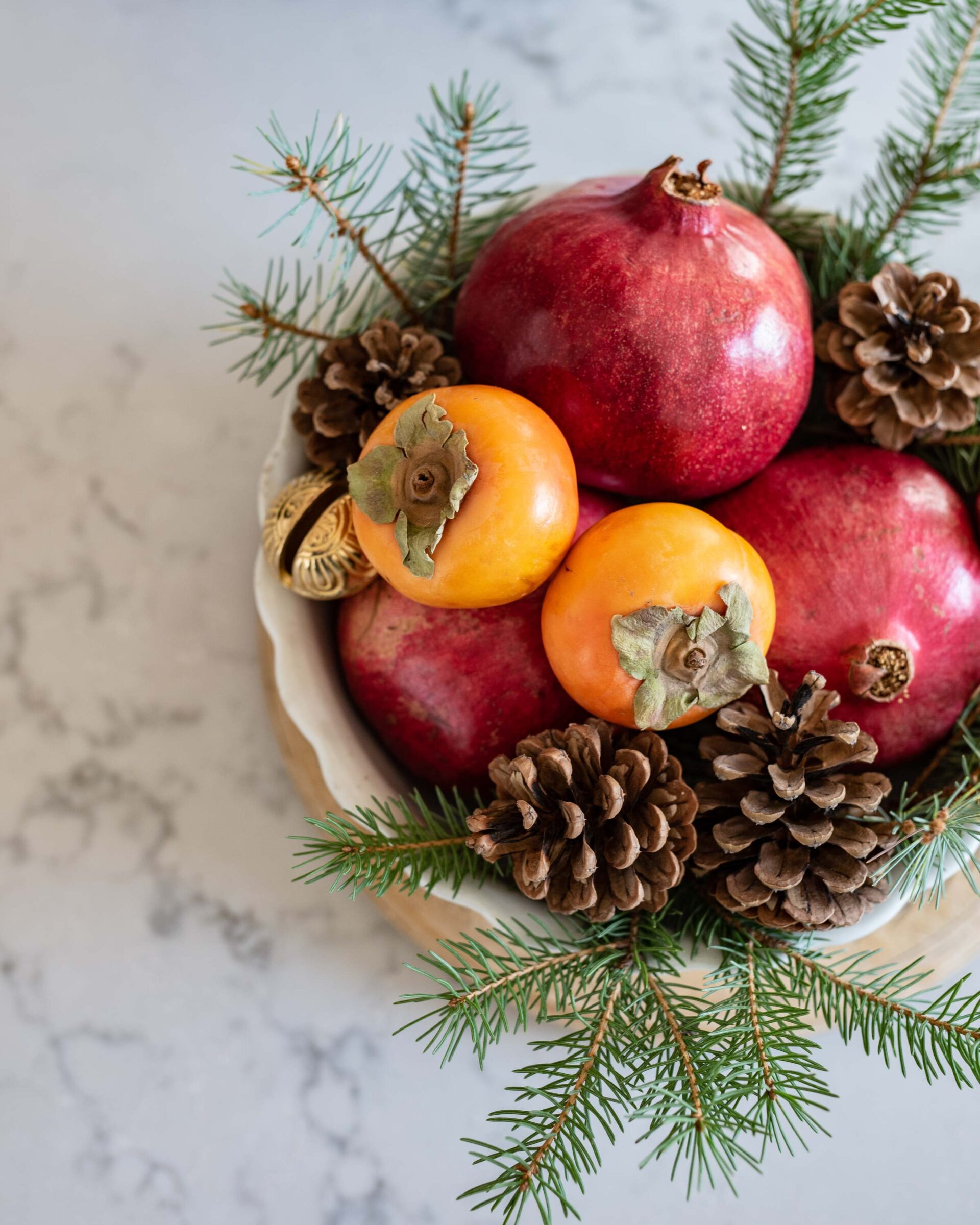 festive-holiday-fruit-bowl-with-pomegranates-and-persimmons-pine-sprigs-and-pinecones-on-marble-surface