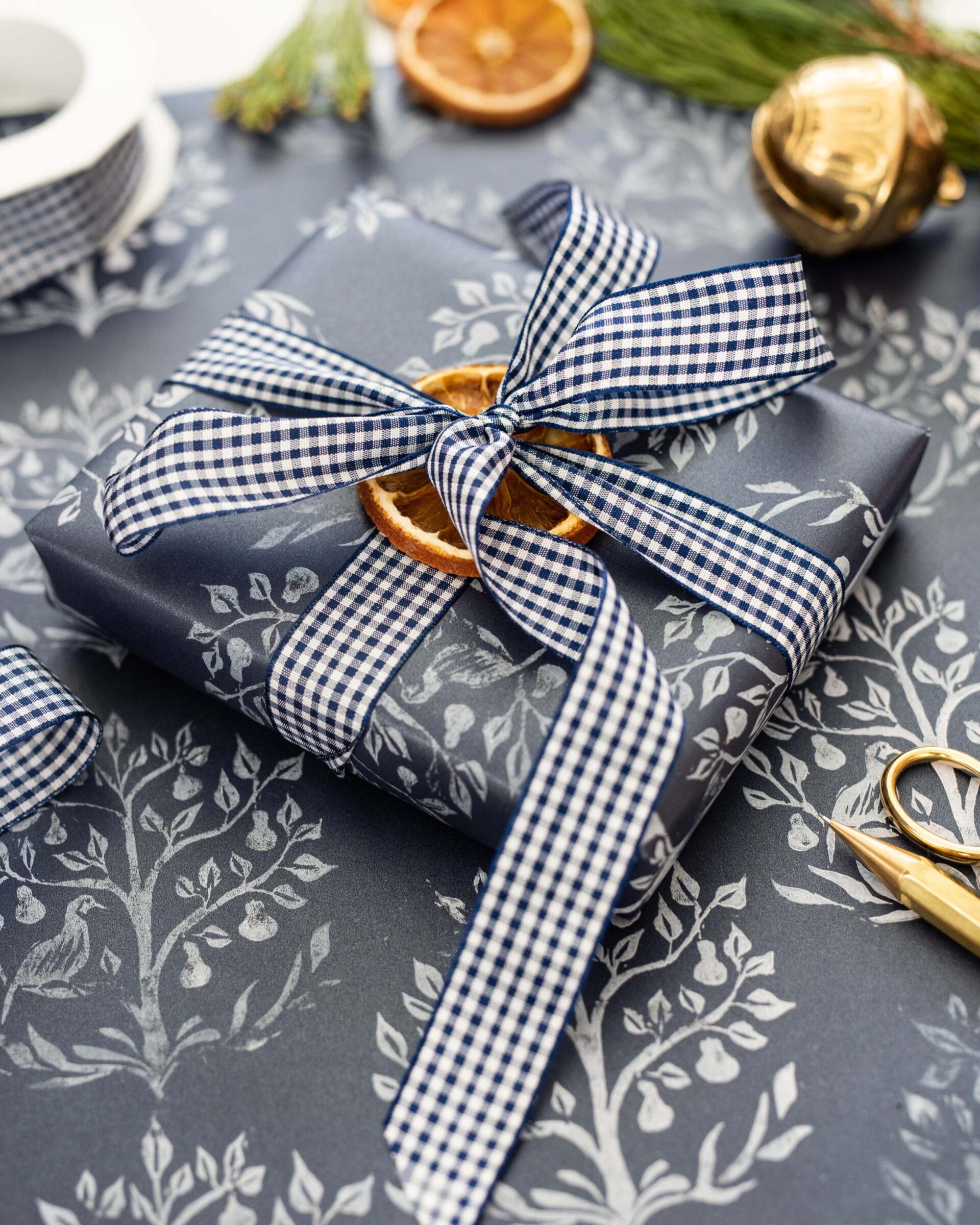 festive-holiday-gift-wrapped-in-blue-gingham-checkered-fabric-with-ribbon-bow-sitting-on-gray-background-with-leaf-pattern