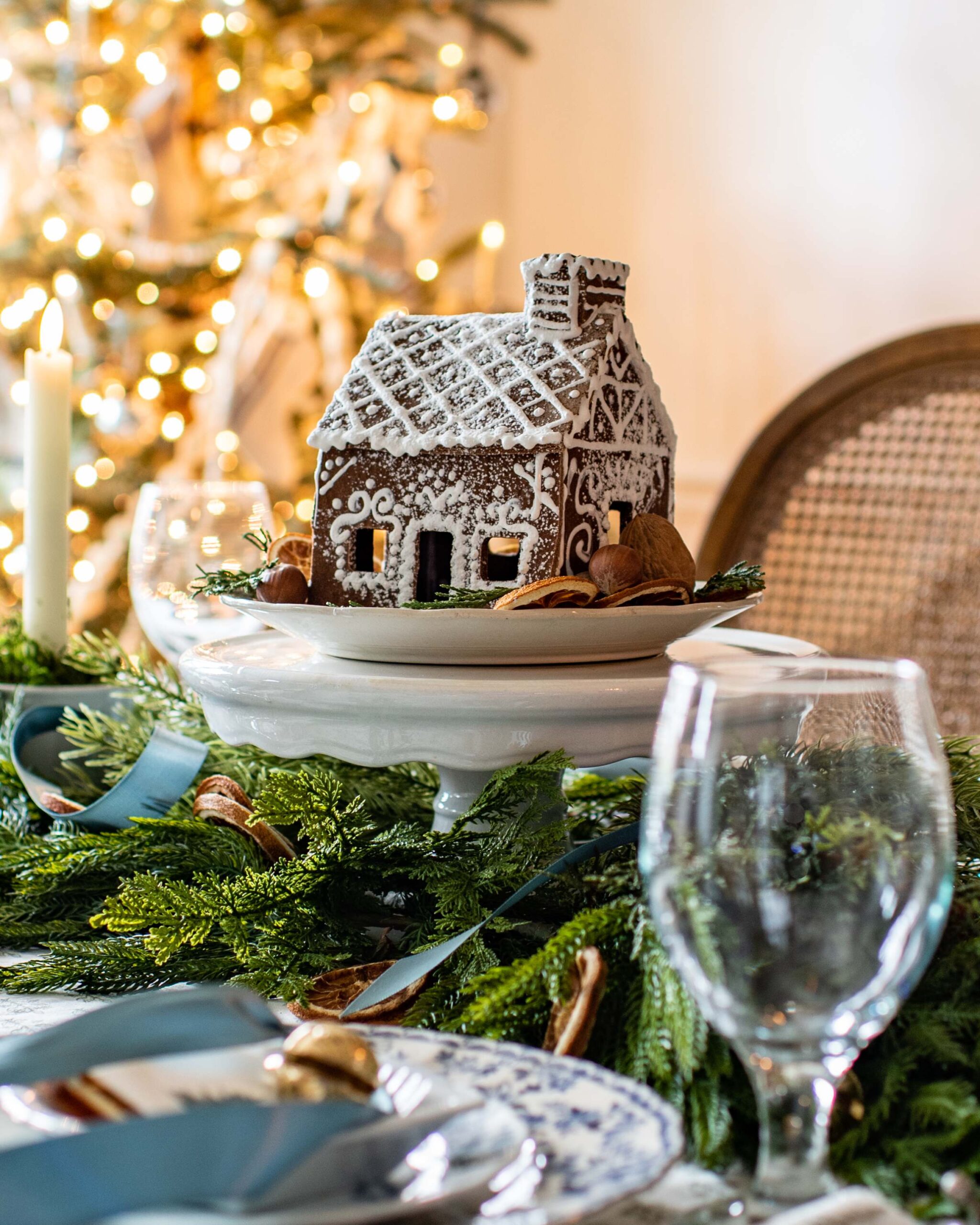 Festive holiday table setting with gingerbread house centerpiece, illuminated Christmas tree in background, blue and white dinnerware, greenery and nuts scattered on wood surface artist reference photo