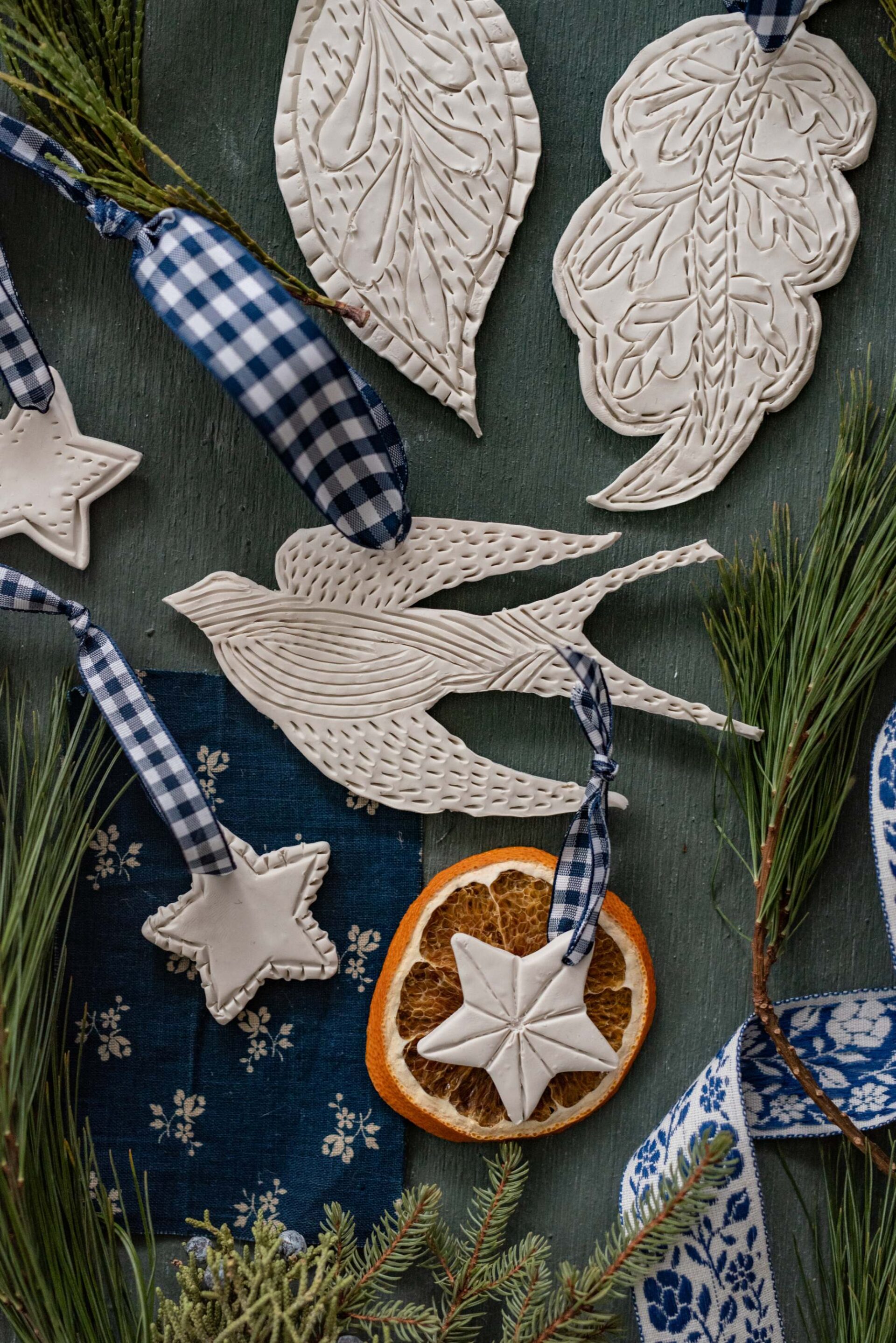 handcrafted-ceramic-christmas-ornaments-textured-white-doves-leaves-and-stars-with-blue-gingham-ribbon-and-dried-orange-slice-on-rustic-green-wood-background