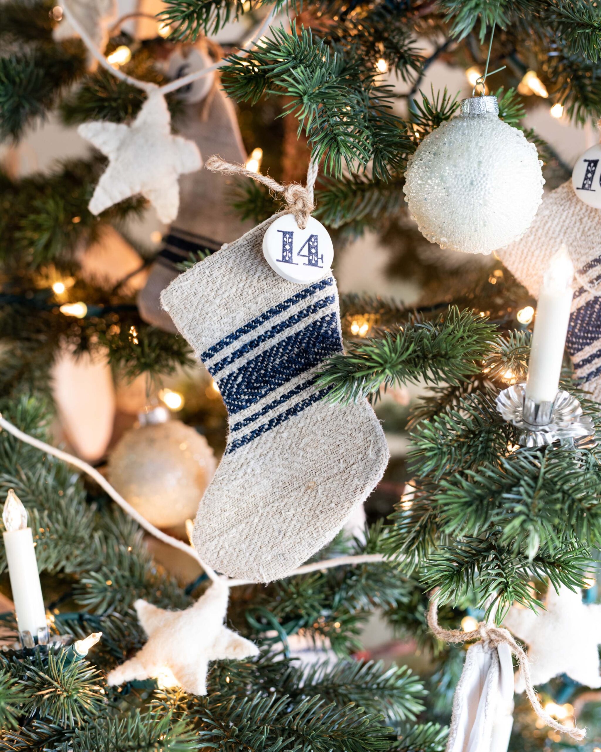 Handmade knitted Christmas stockings with blue stripes and personalized initial tags hanging from Christmas tree branches adorned with lights and ornaments artist reference photo