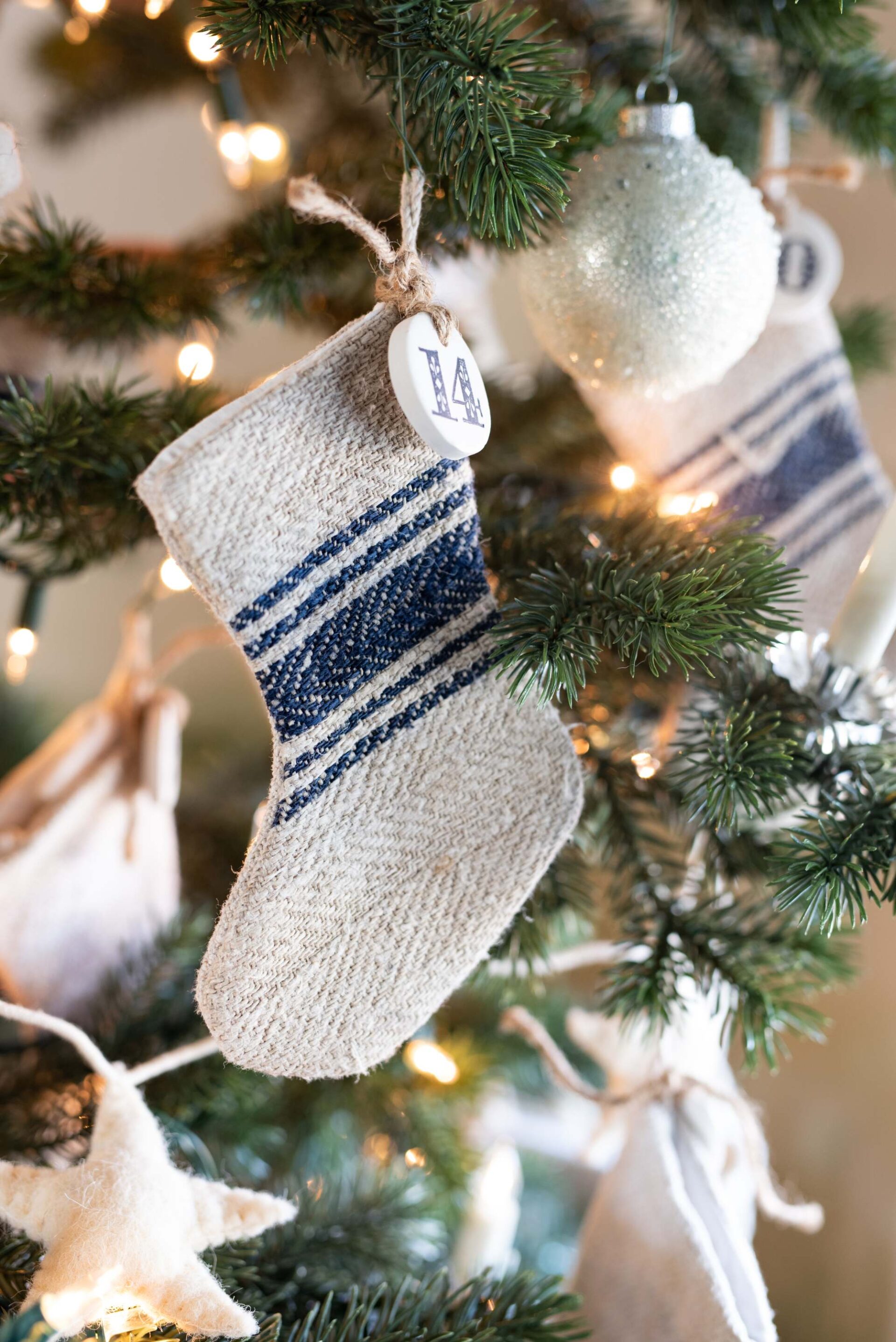 knitted-christmas-stocking-with-beige-body-and-blue-striped-cuff-hanging-from-christmas-tree-branch-with-personalized-initial-tag