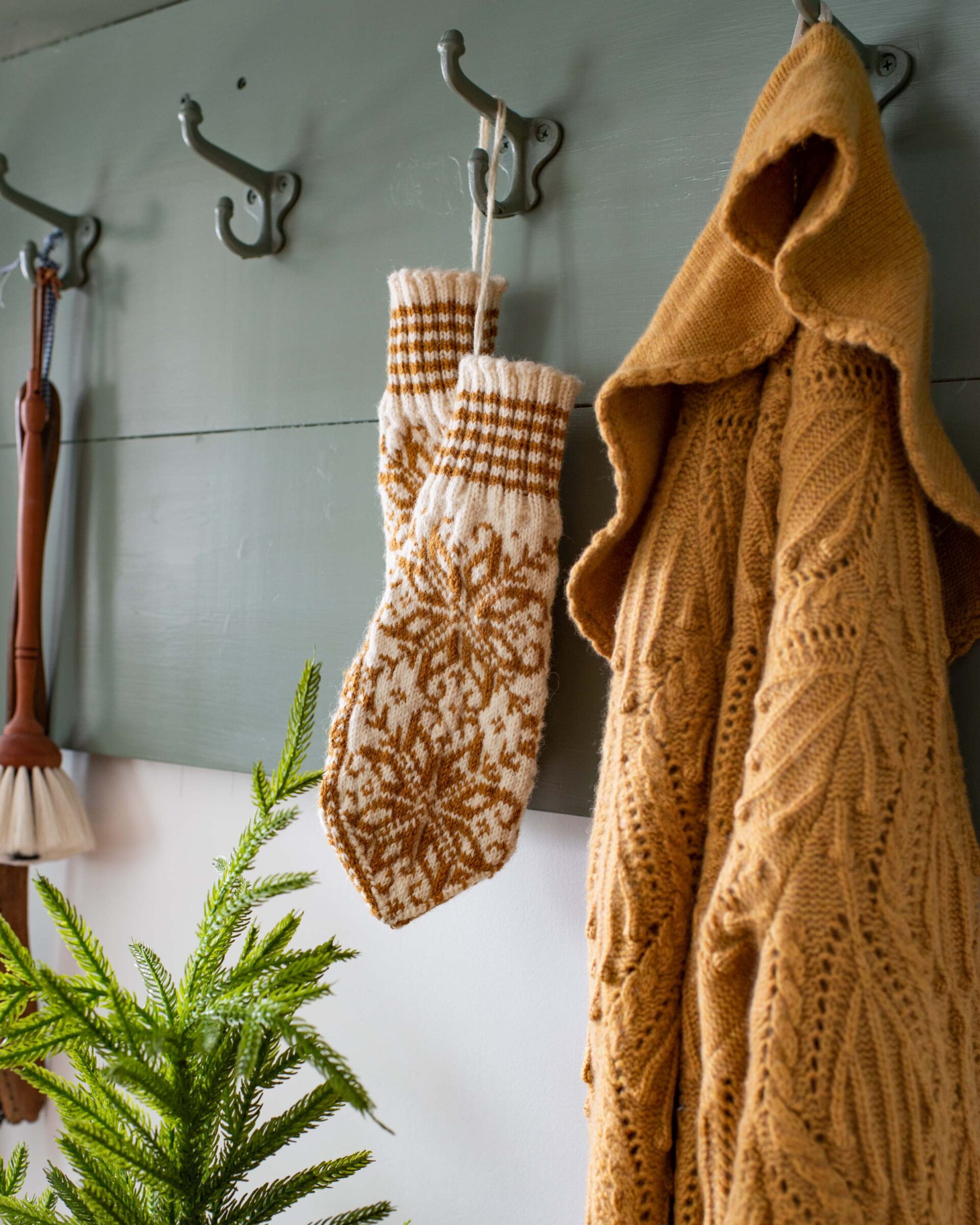 knitted-winter-accessories-including-white-and-brown-patterned-mittens-and-a-tan-cable-knit-hooded-sweater-hanging-from-hooks-against-a-light-blue-wall-with-an-evergreen-branch-adding-a-festive-holid