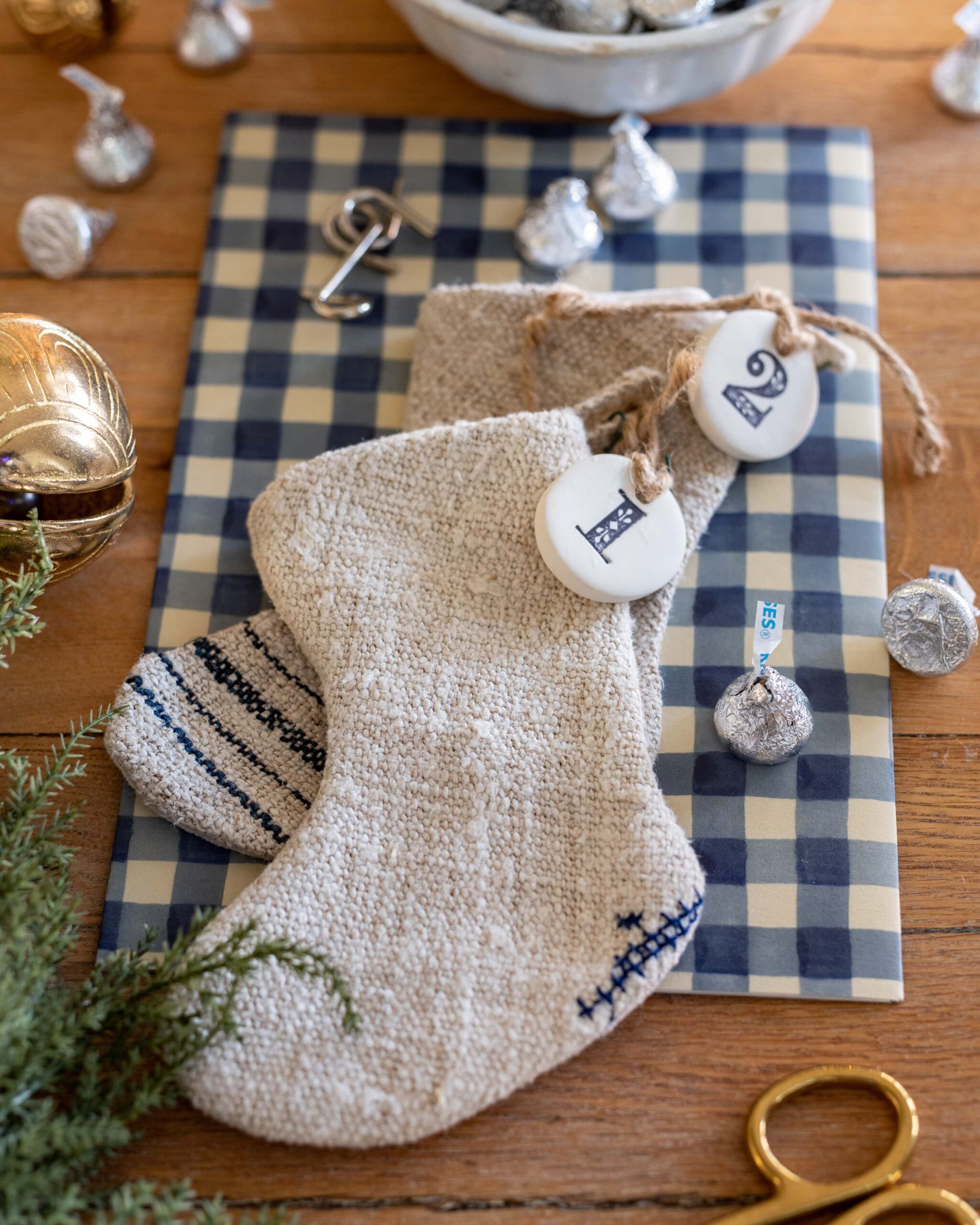 Knitted wool Christmas stocking with blue checked pattern, personalized initial tags, and chocolate Hershey’s Kisses scattered around on wood table with gold accents artist reference photo