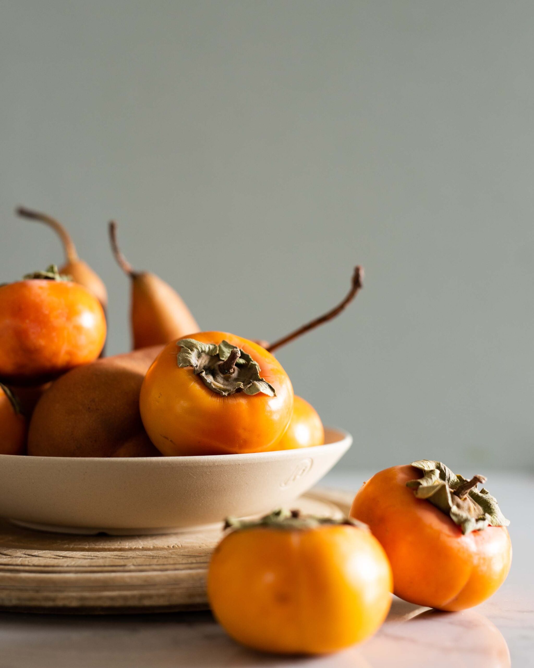 Persimmons in a white bowl, some sliced open showing their textured flesh, garnished with leafy stems against a light gray background artist reference photo