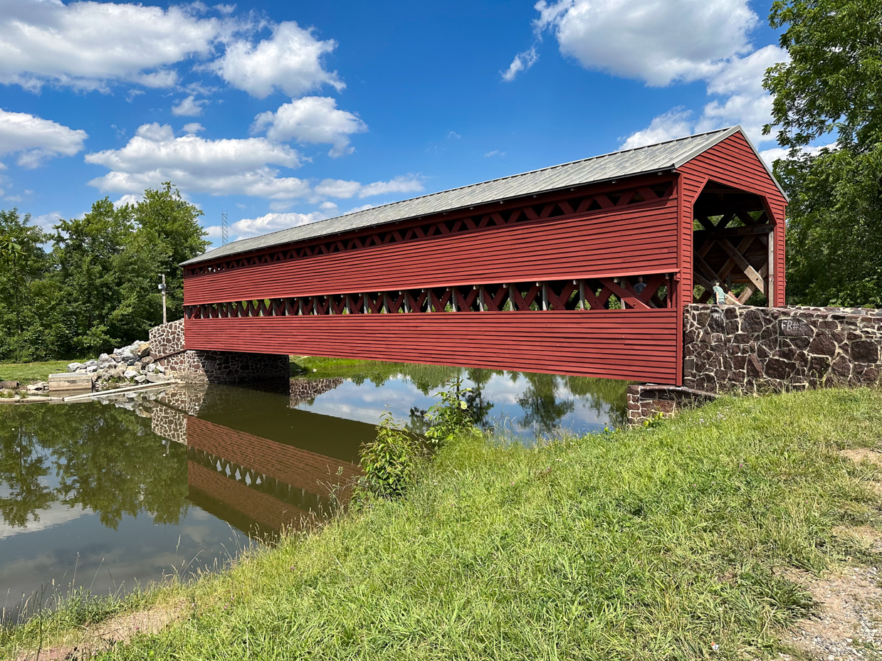 red-covered-bridge-spanning-river-scenic-rural-landscape-fluffy-white-clouds-blue-sky-stone-abutments-reflection-water-summer-greenery-historic-architecture-americana-royalty-free-artist-reference-pho