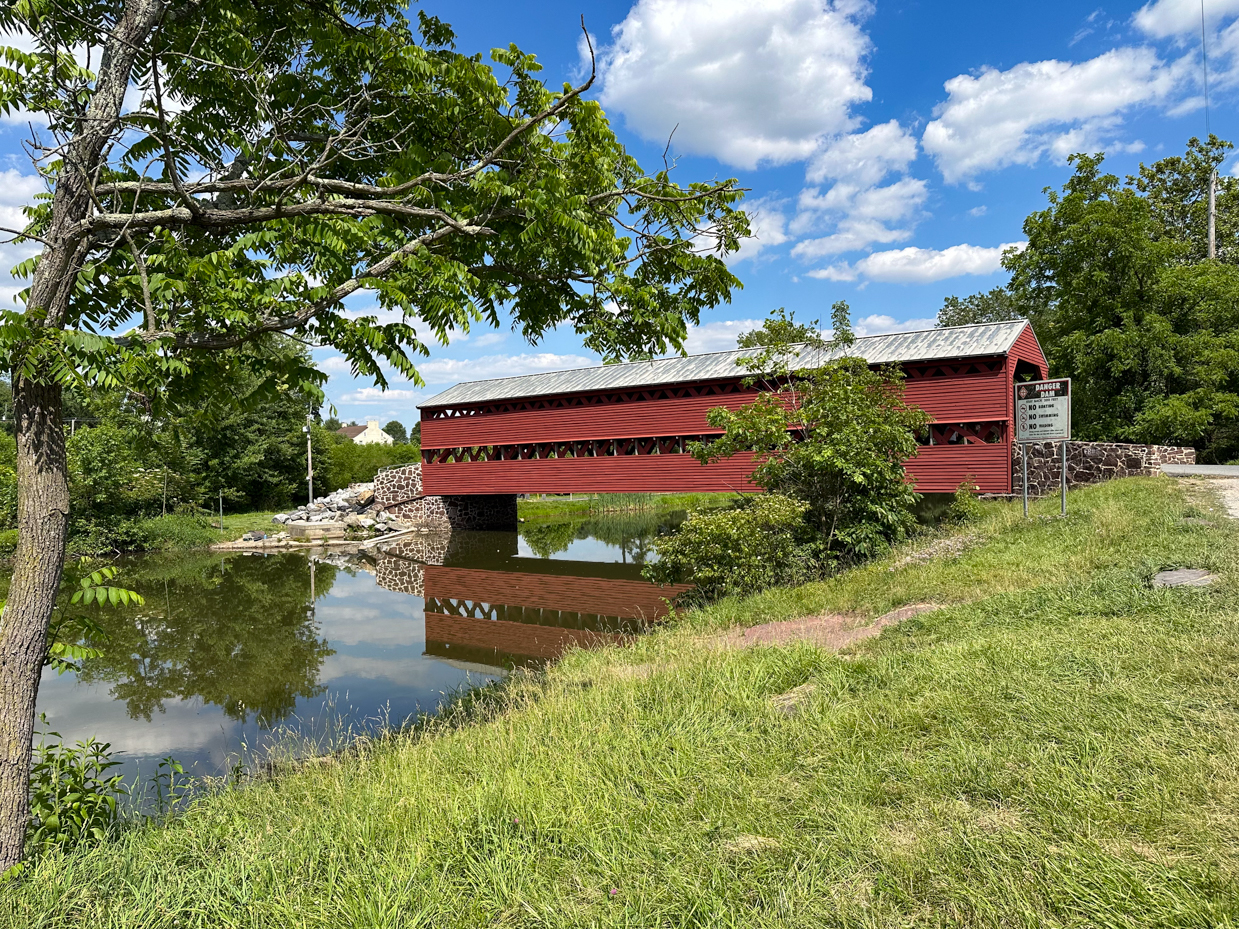 rural-covered-bridge-scenic-landscape-summer-day-vibrant-red-wooden-structure-spanning-stream-lush-green-foliage-blue-sky-puffy-clouds-stone-foundations-peaceful-countryside-scene-water-reflection-his