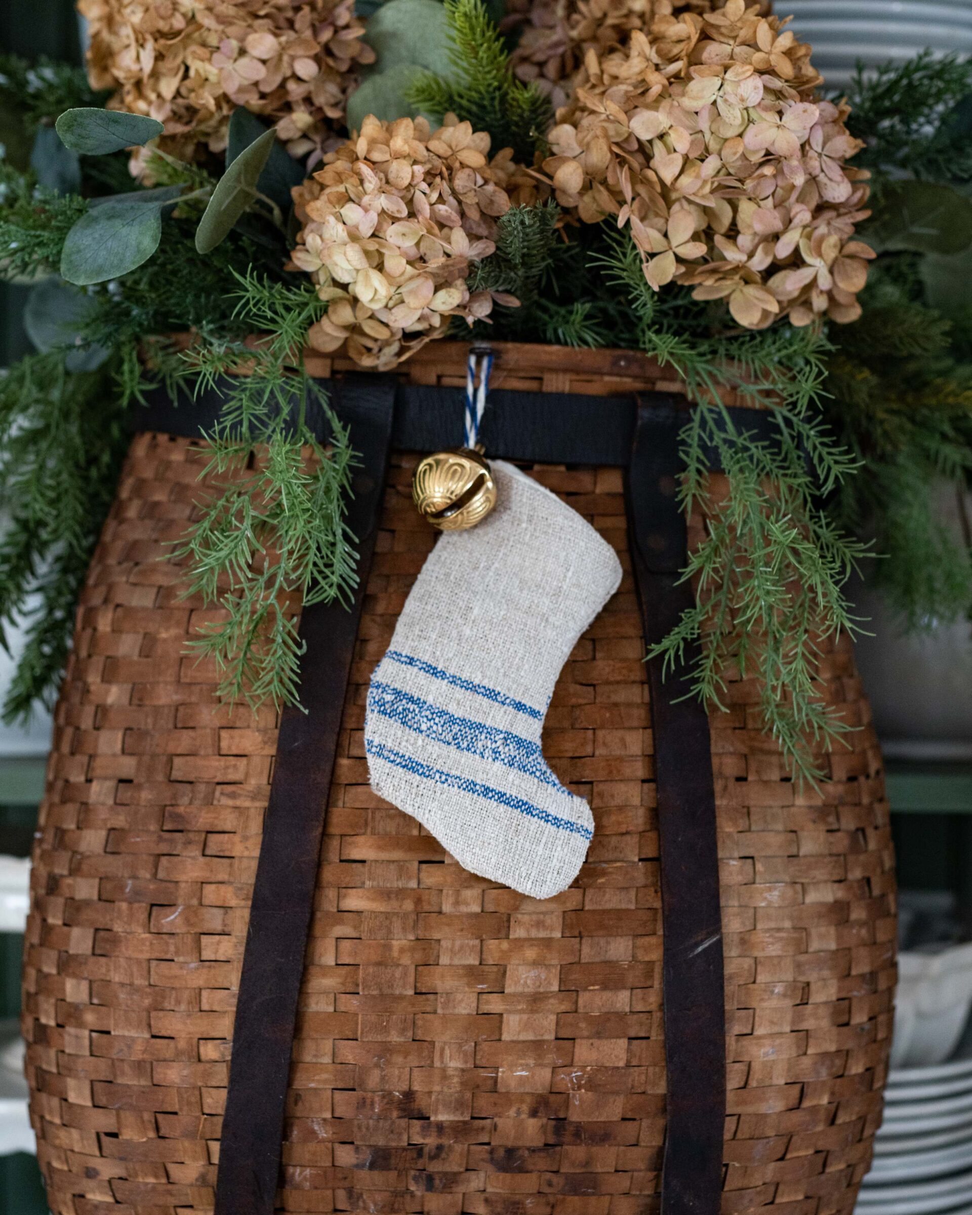 rustic-farmhouse-christmas-woven-stocking-with-blue-stripe-detail-hangs-from-gold-jingle-bell-on-textured-woven-basket-filled-with-dried-hydrangea-flowers-and-fresh-greenery