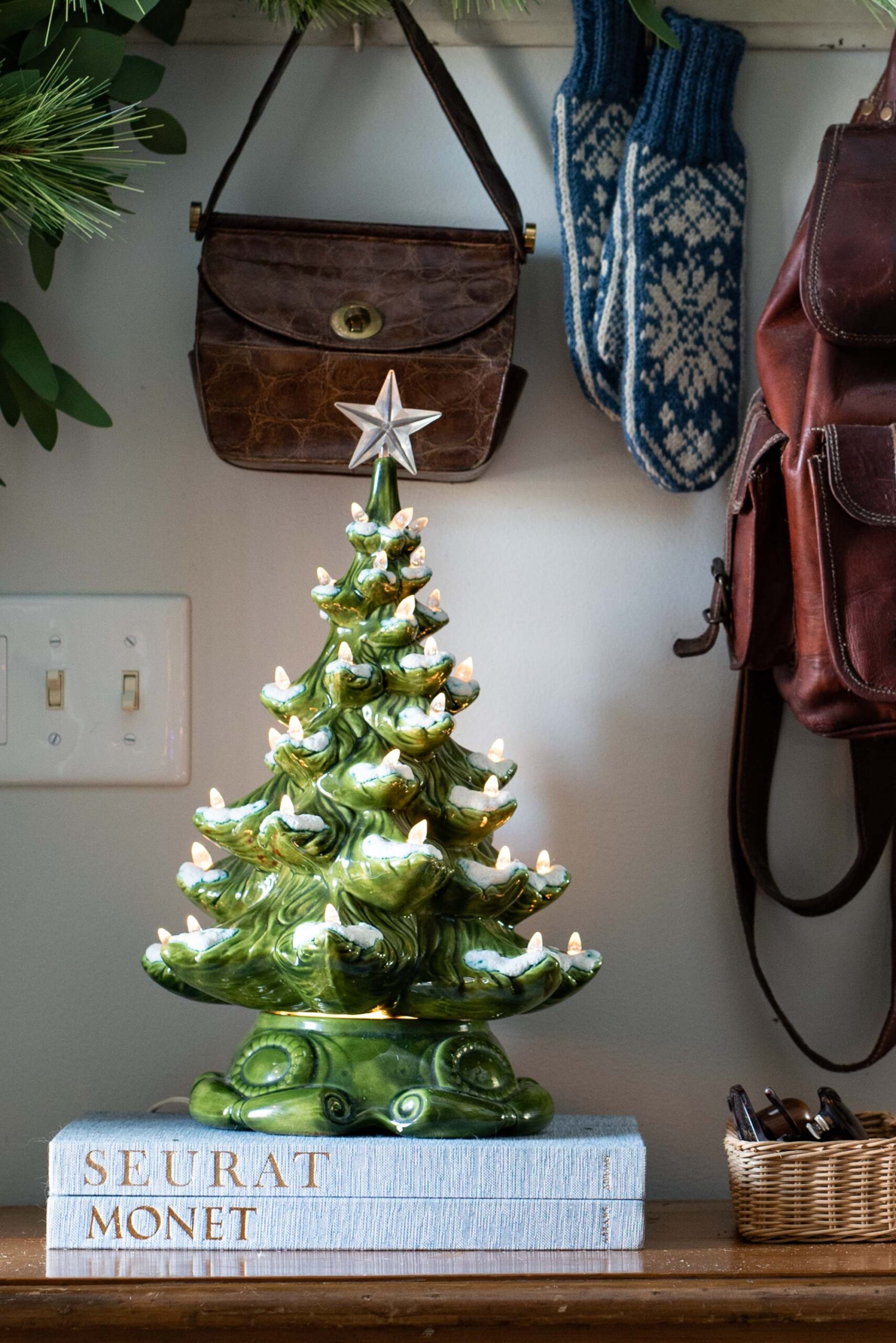 Vintage ceramic Christmas tree with lit candles displayed on books labeled “Seurat Monet,” with leather satchel and winter accessories hanging on wall artist reference photo