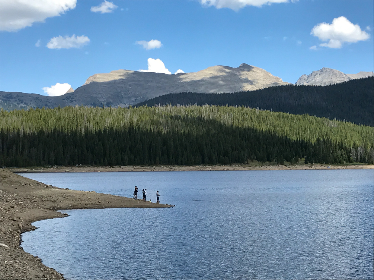 hikers-on-rocky-shoreline-of-alpine-lake-with-pine-forest-and-snow-capped-peaks-royalty-free-photo