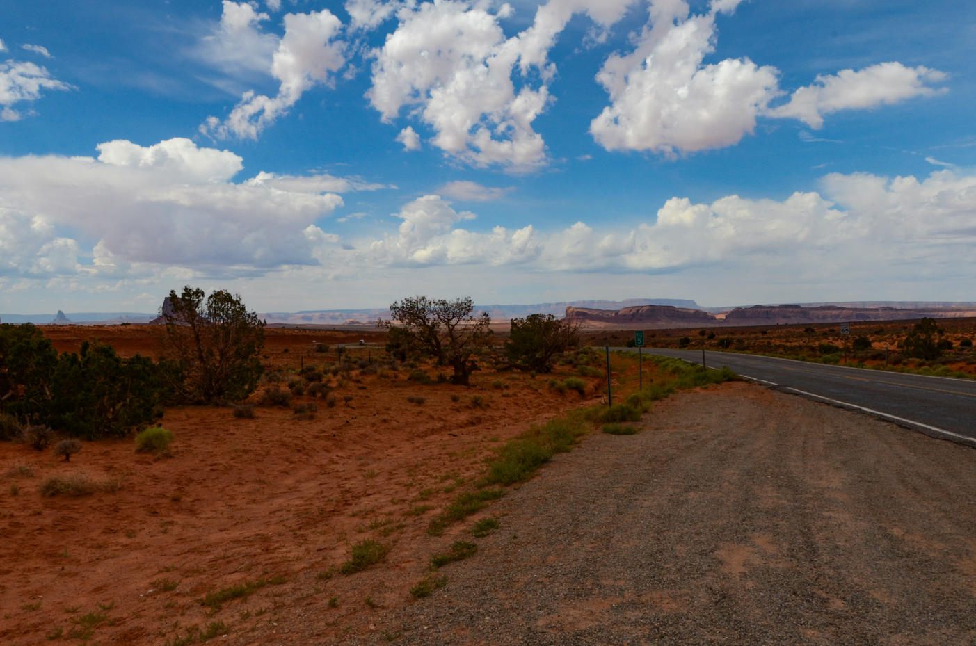 winding-desert-highway-through-scrubland-towards-distant-monument-valley-mesas-under-puffy-clouds