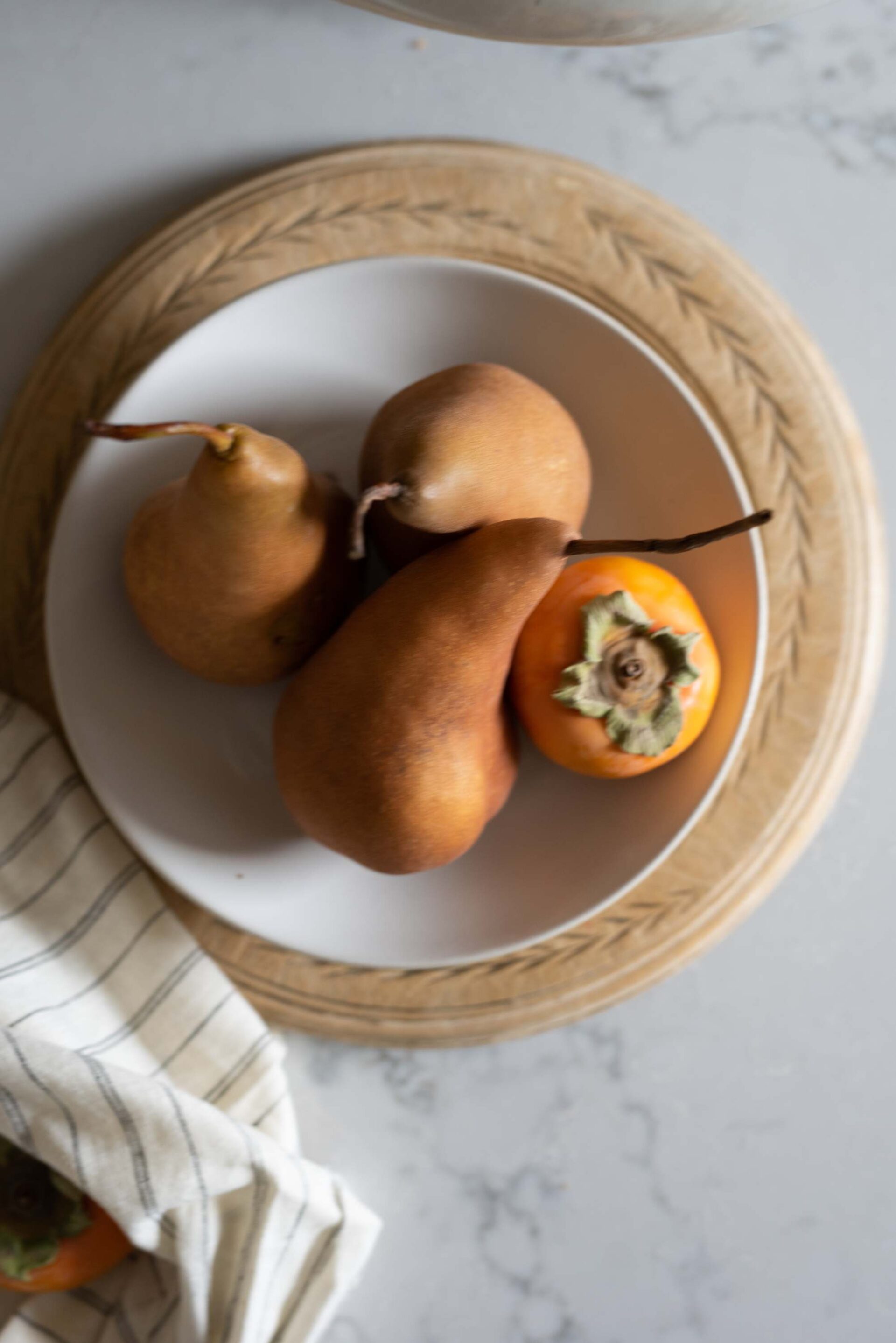 still-life-of-three-bosc-pears-and-a-persimmon-on-a-white-plate-placed-on-a-wooden-charger-with-leaf-patterns-accompanied-by-a-striped-cloth-on-a-marble-surface