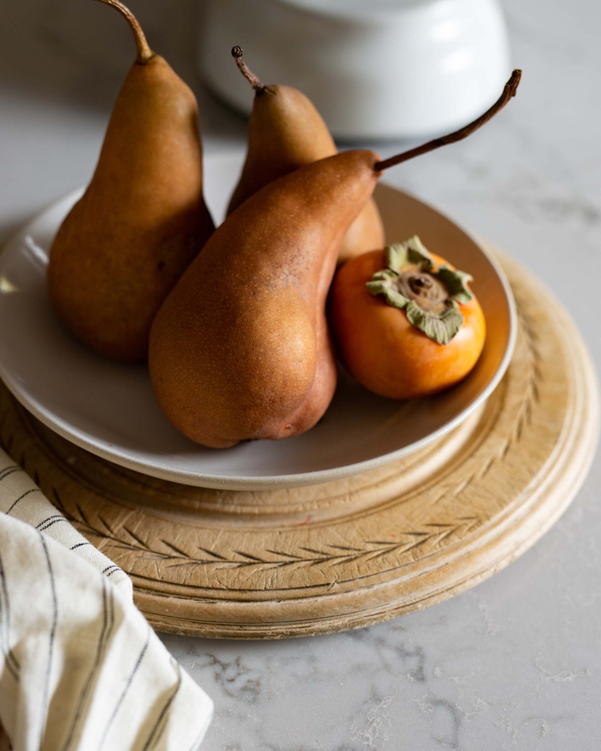 still-life-of-three-bosc-pears-and-a-persimmon-on-a-white-plate-set-on-an-ornate-wooden-charger-with-a-striped-cloth-and-marble-countertop-in-soft-light