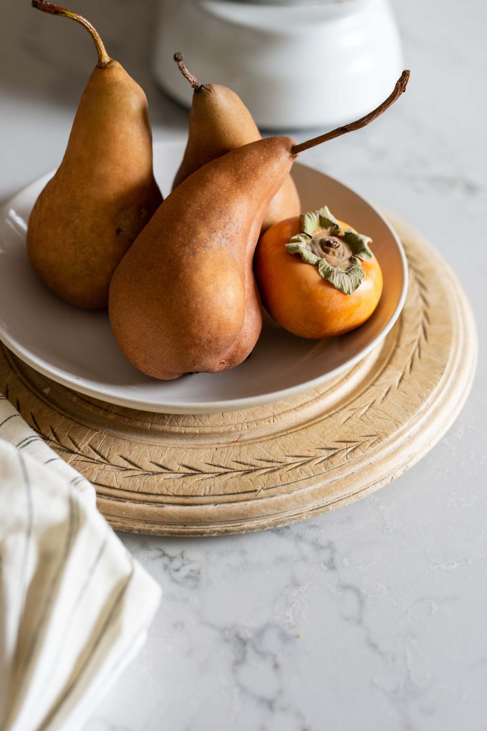 Three Bosc pears and a persimmon on a white plate, arranged on a wooden charger with a striped cloth on a marble surface artist reference photo