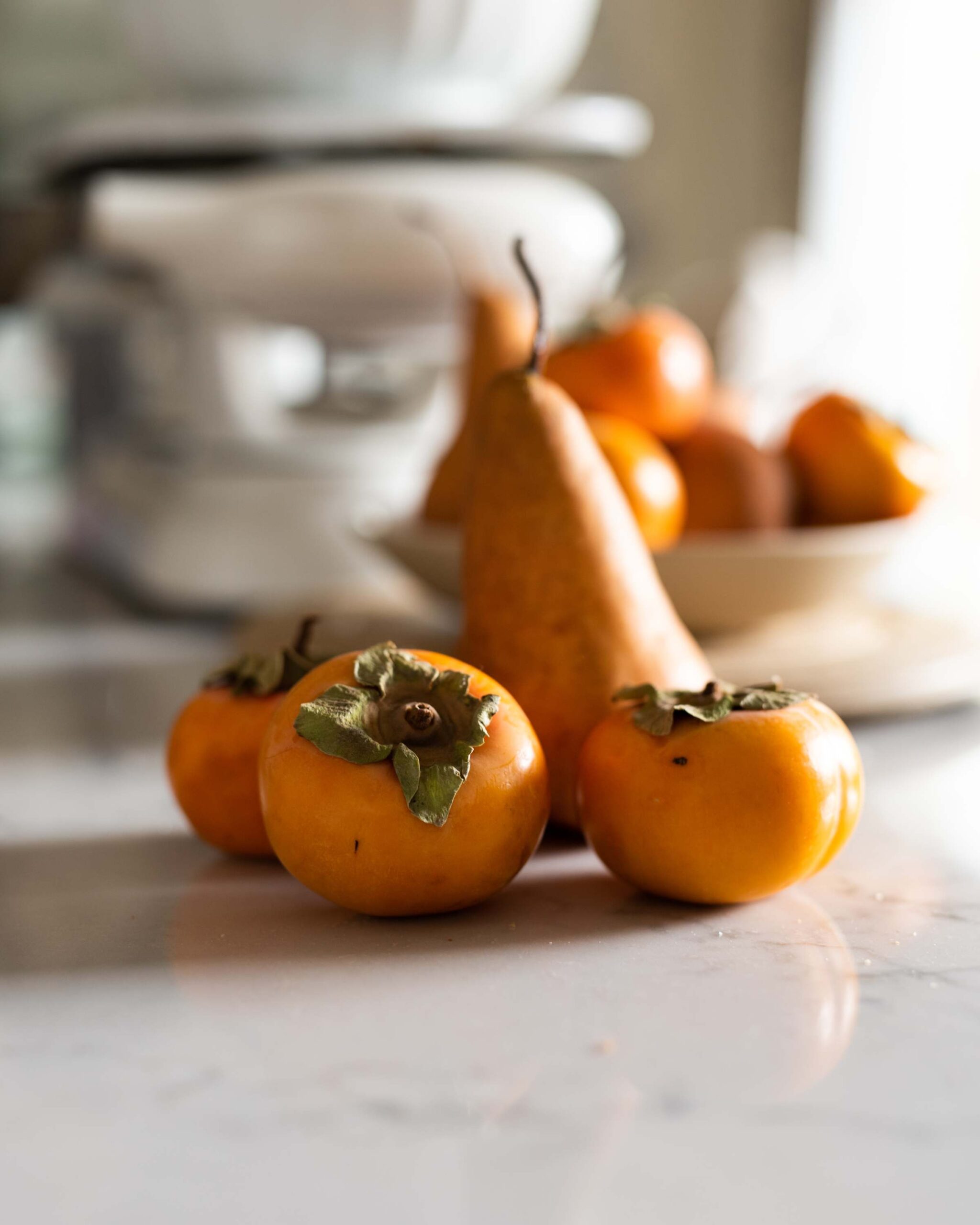 Three persimmons and a Bosc pear on a reflective marble surface, with blurred background elements including a bowl and scale artist reference photo