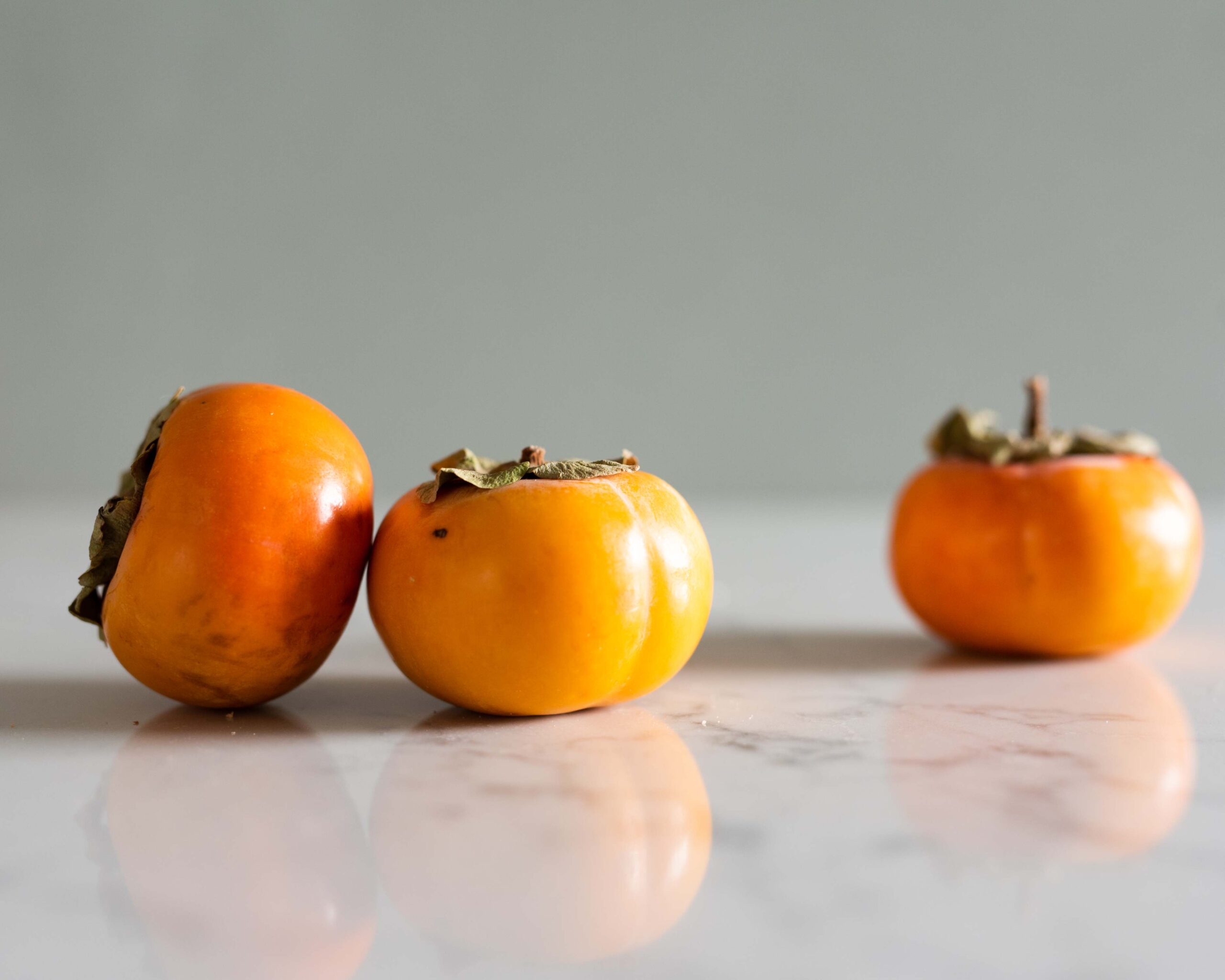 Three persimmons arranged on a reflective marble surface, their orange hues contrasting against a soft, neutral background artist reference photo