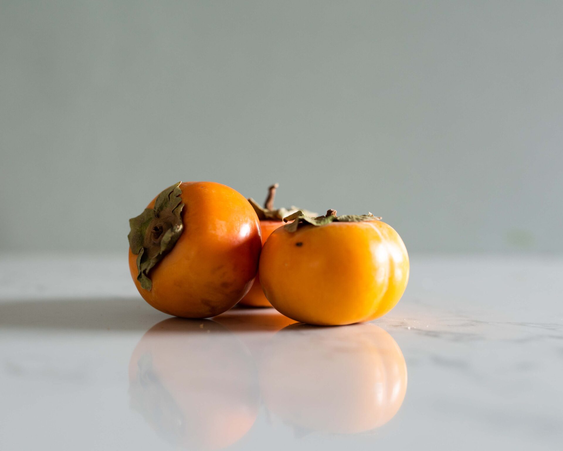 three-persimmons-arranged-on-a-reflective-marble-surface-with-a-soft-green-background-emphasizing-their-vibrant-orange-color