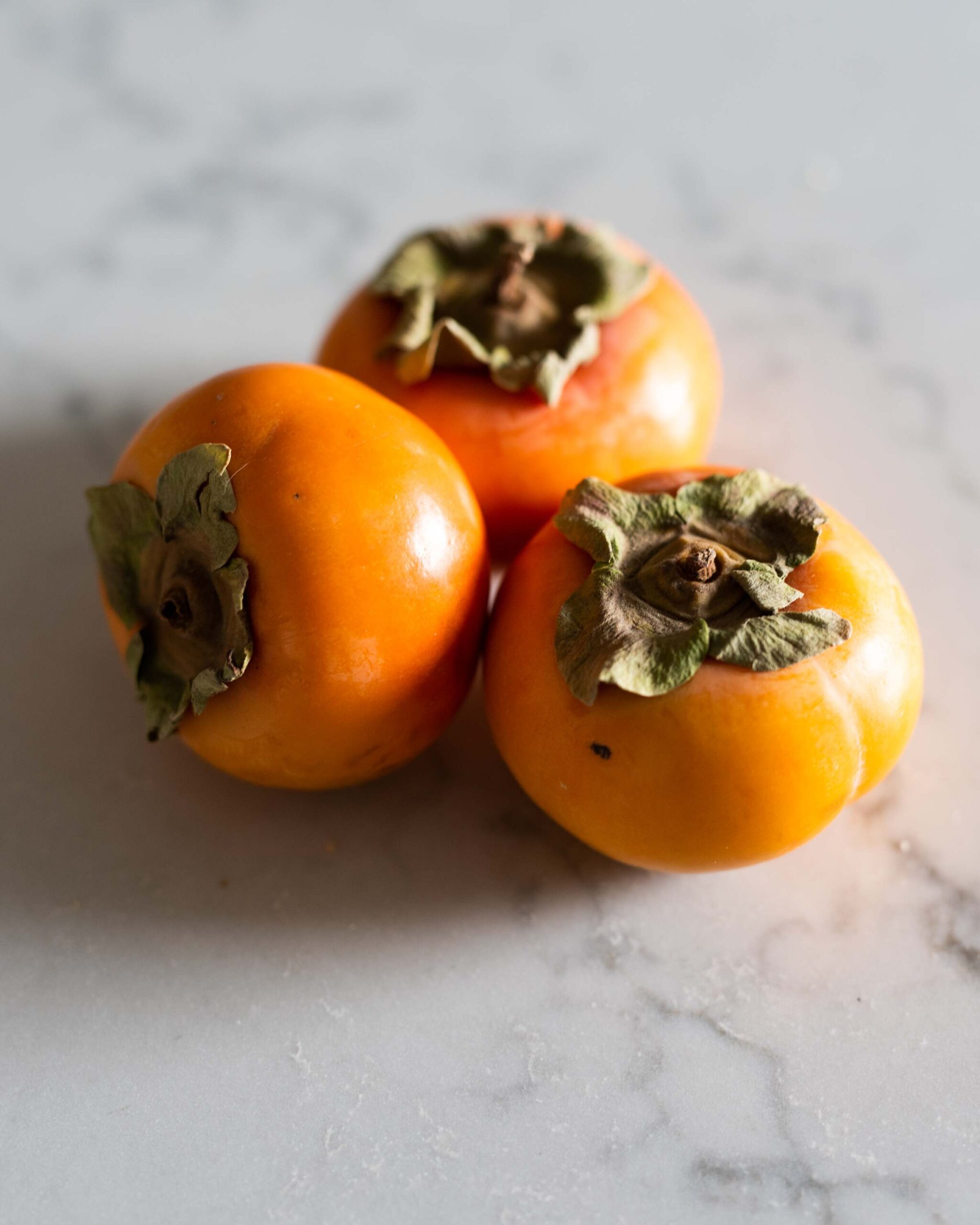 Three persimmons resting on a light marble surface, showcasing their vibrant orange tones and green leafy tops artist reference photo