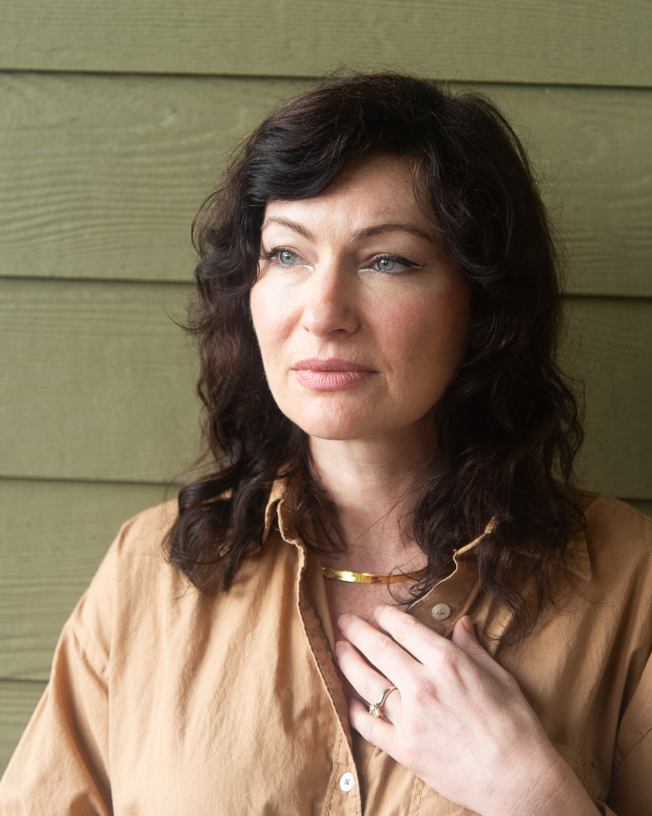 woman-with-dark-wavy-hair-and-gold-necklace-in-tan-shirt-touching-collar