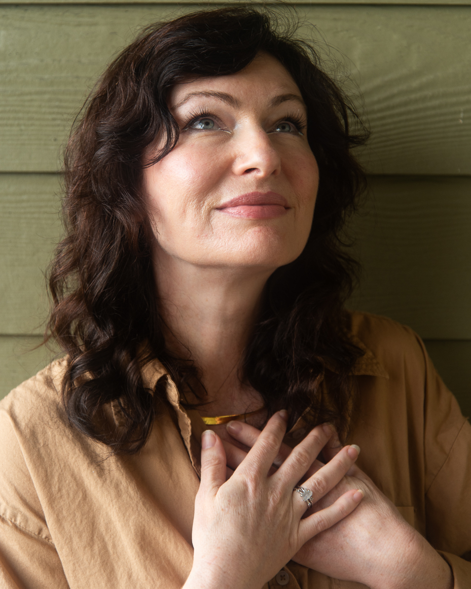 woman-with-wavy-dark-hair-in-tan-shirt-looking-up-with-hands-over-heart-and-soft-smile