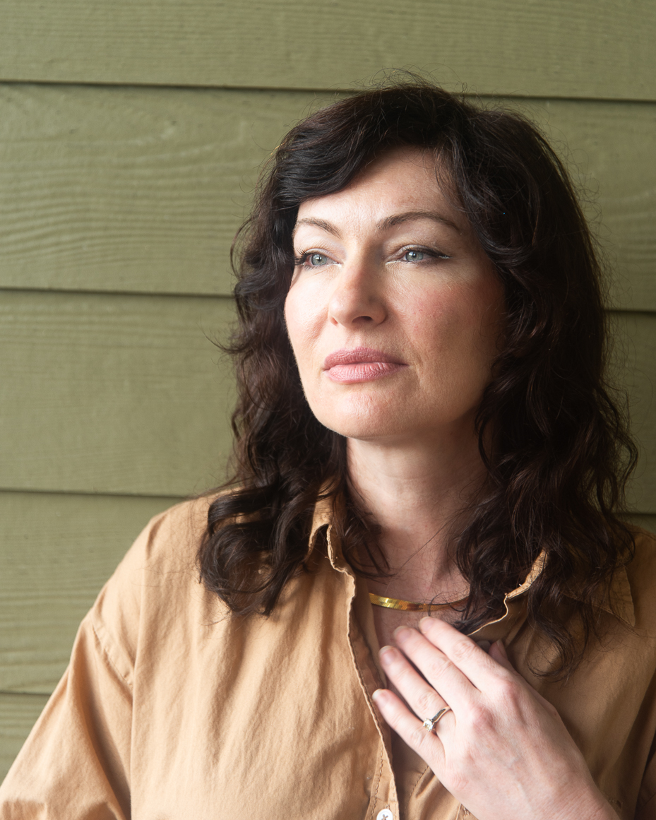 woman-with-wavy-dark-hair-touching-necklace-in-tan-button-up-shirt