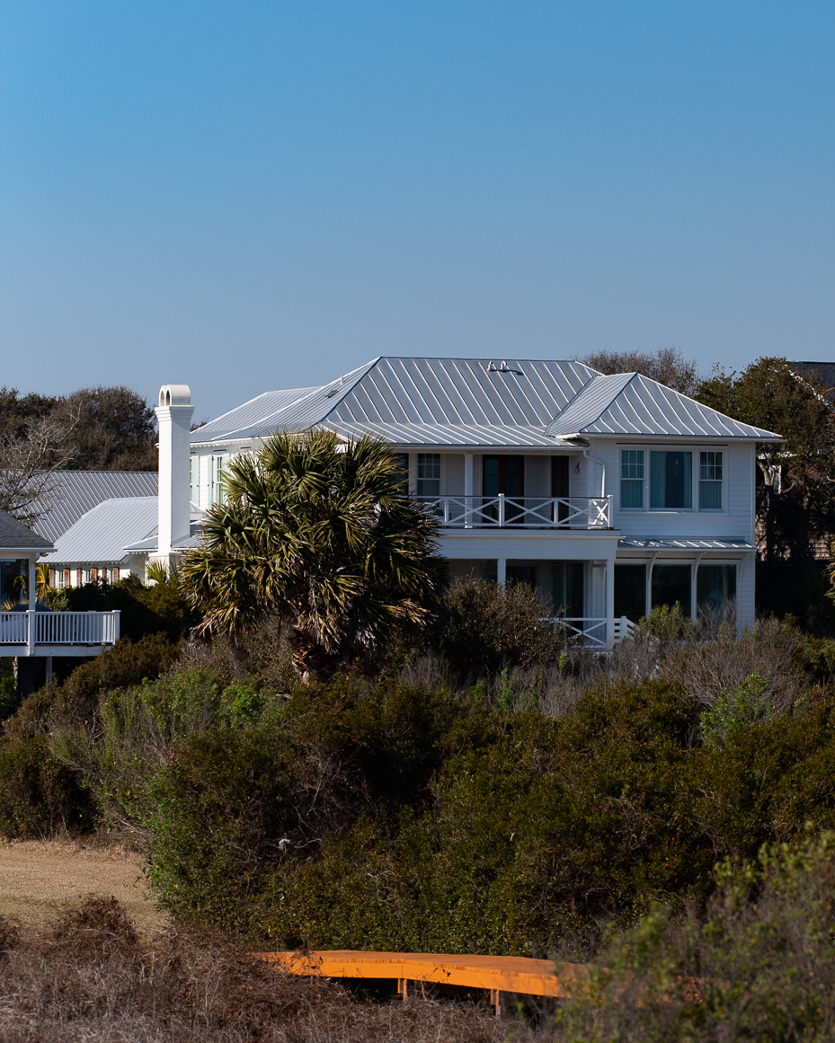 white-beach-house-with-metal-roof-and-wraparound-porch-behind-coastal-greenery